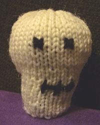 A knitted off-white small toy skull with the eyes, nose and teeth outlined in black yarn